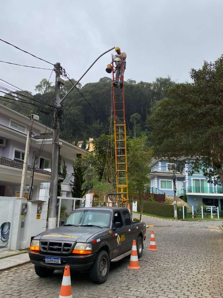Profissional da Nerel Elétrica sobre escada realizando manutenção de luminária de iluminação pública em rua residencial, com caminhonete da empresa e cones de sinalização