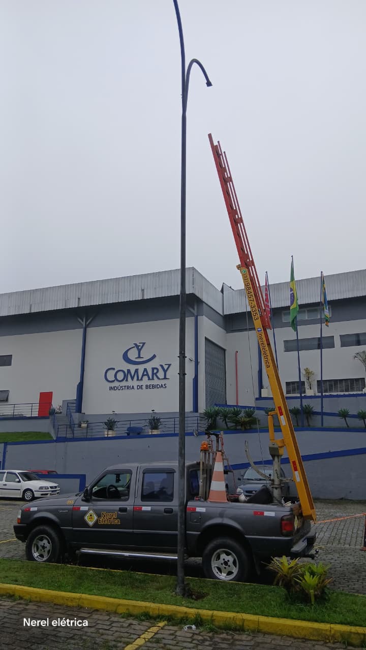 Caminhonete e escada de trabalho da Nerel Elétrica posicionadas para manutenção de poste de iluminação em área industrial
